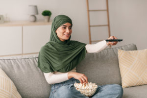 person with a hijab on with a TV remote in their hand and a bowl of popcorn in their lap sitting on a couch